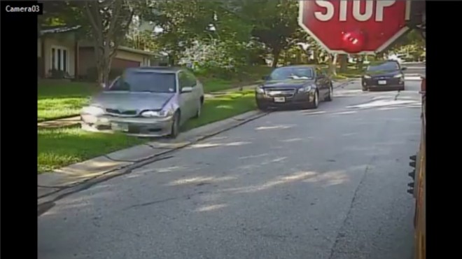 The silver sedan in this video screen is cutting through the lawn and around a school bus.