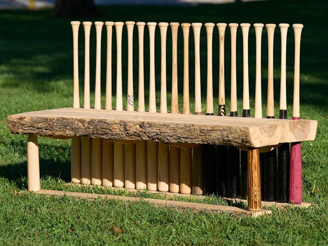A bench where the back is made from baseball bats sits in a park.