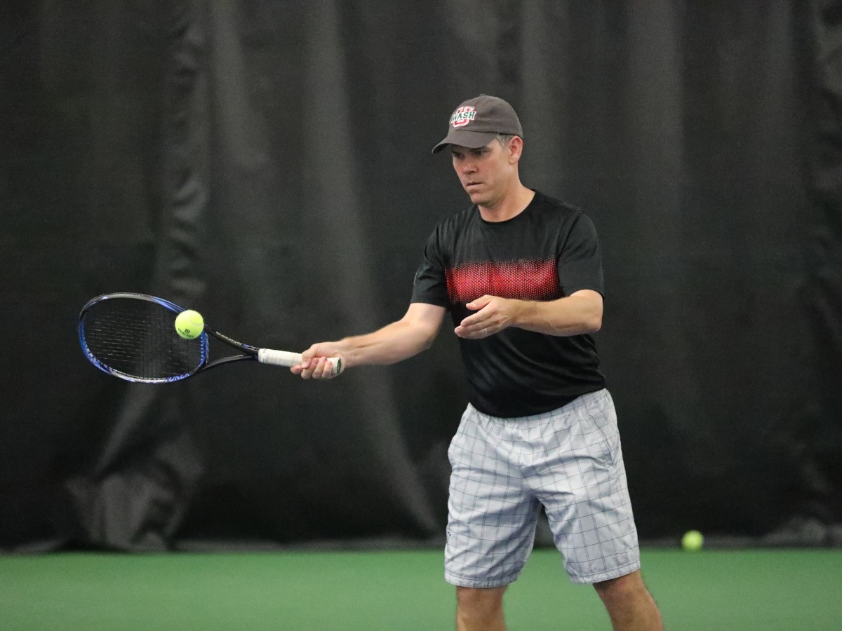 Coach Roger Follmer swings a tennis racket at a ball.