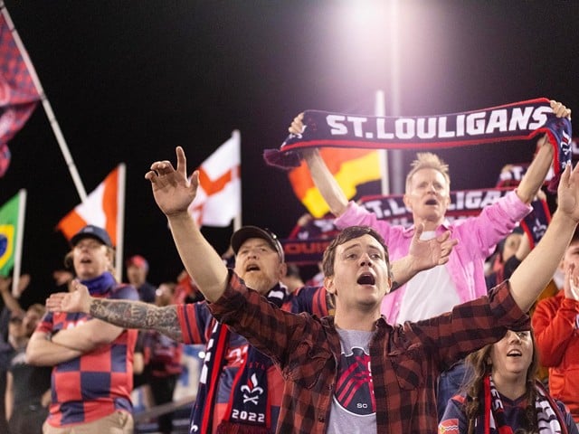 Soccer fans raise their hands, hold a "St. Louiligans" scarf and fly flags of different countries as they cheer from the stands.