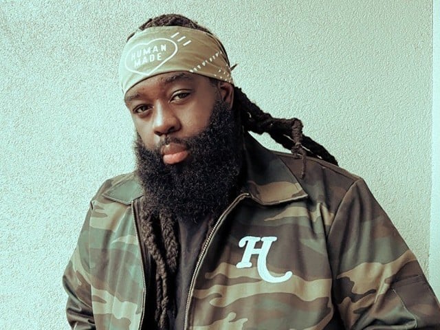Jas Bell looks at the camera wearing a bandana and a camo jacket with the letter "H."