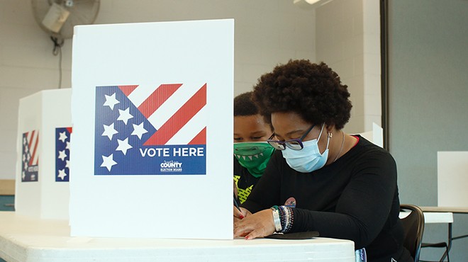 A woman with a blue surgical mask fills out a ballot behind a white "vote here" sign as her son watches from behind.