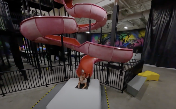 A kid slides down a red slide on a mat with basketball hoops in the background.