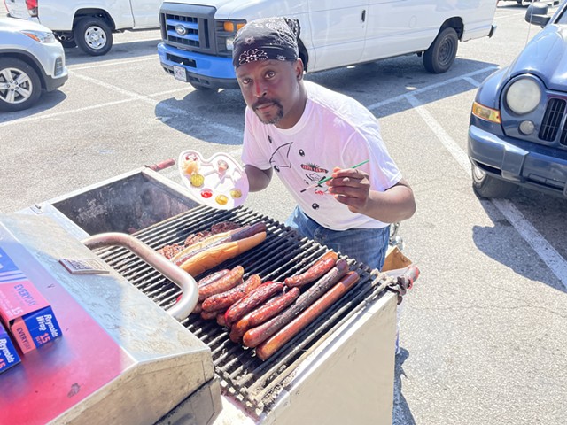 Larry Lunceford leans in front of his hot dog cart. He holds a palette in one hand with mustard, ketchup, pickles and more. In his other hand, he holds a paintbrush. In front of him is food on a grill, including long hot dogs, short dogs and burgers. He's standing in a parking lot.