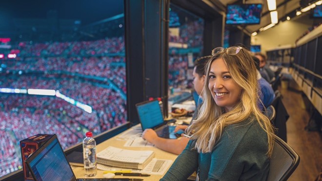 Katie Woo, St. Louis Cardinals beat reporter sits in the press box with a packed stadium crowd behind her.