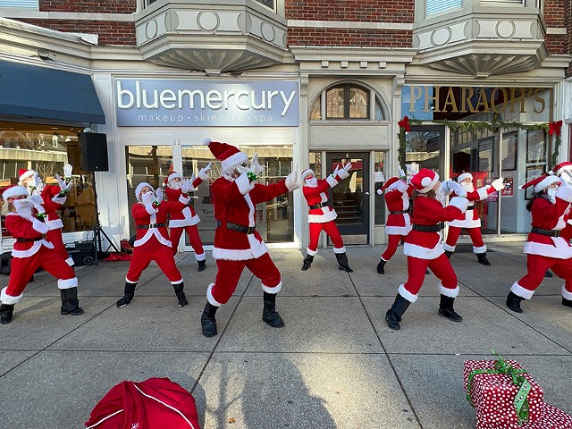 People dressed up in Santa costumes dance on the sidewalk.