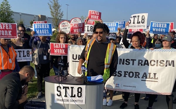 Union organizer J. Lopez speaks at a rands with a crowd of people behind him. They hold signs such as "STL8 Organizing Committee," "Safer Work" and "Higher Wages."