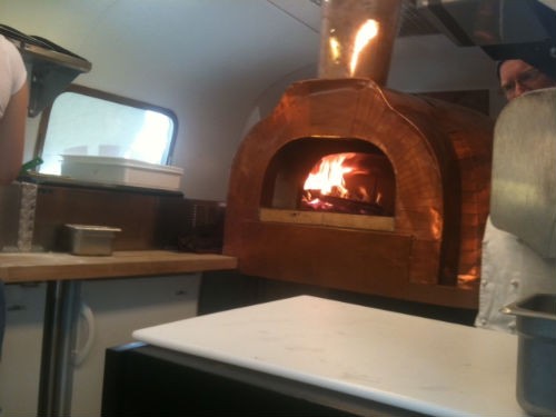 Chef Erik Jacobs peeks from behind Wanderlust Pizza's wood-fired oven inside an Airstream trailer. - Robin Wheeler
