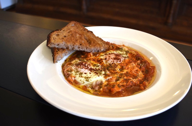 Shakshuka made with a base of tomato and pepper stew, poached eggs, harissa, feta, marinated cucumbers and herbs served with sourdough flatbread. - Liz Miller