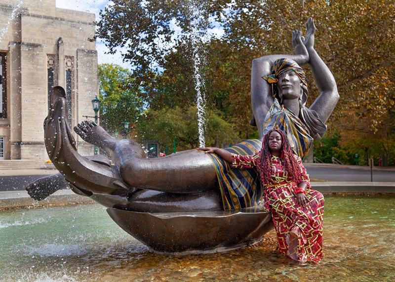 The artist leans against a large bronze statue of a woman dressed in a multicolor fabric.