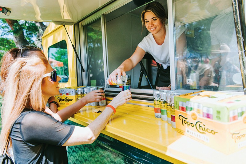A woman smiles as she leans out of a food truck and hands a customer an alcoholic seltzer.