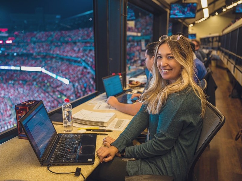 Katie Woo, St. Louis Cardinals beat reporter sits in the press box with a packed stadium crowd behind her.
