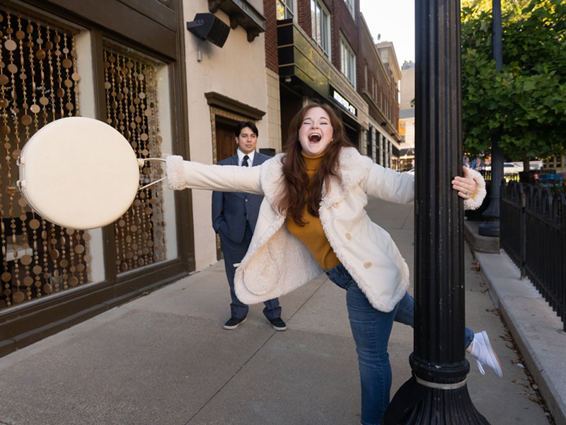 A woman swings joyfully on a lamppost while a man looks on.