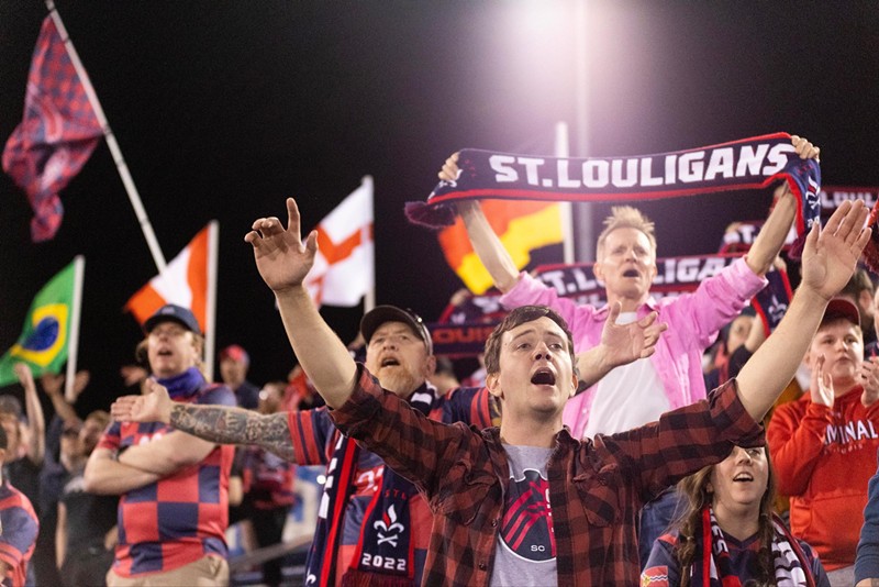Soccer fans raise their hands, hold a "St. Louiligans" scarf and fly flags of different countries as they cheer from the stands.