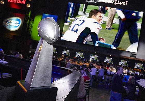 The silver Lombardi Trophy sits on a ledge with a large TV showing Tom Brady in the background of a restaurant.