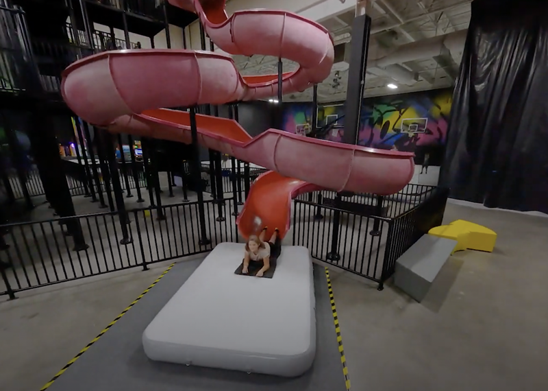 A kid slides down a red slide on a mat with basketball hoops in the background.