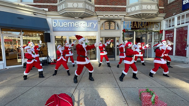People dressed up in Santa costumes dance on the sidewalk.