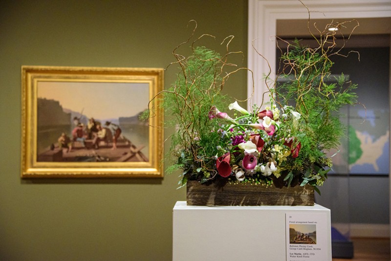 A flower display on top of a white stand in front of a picture.