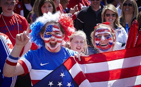 Two United States soccer fans cheer in red, white and blue facepaint while holding an American flag.